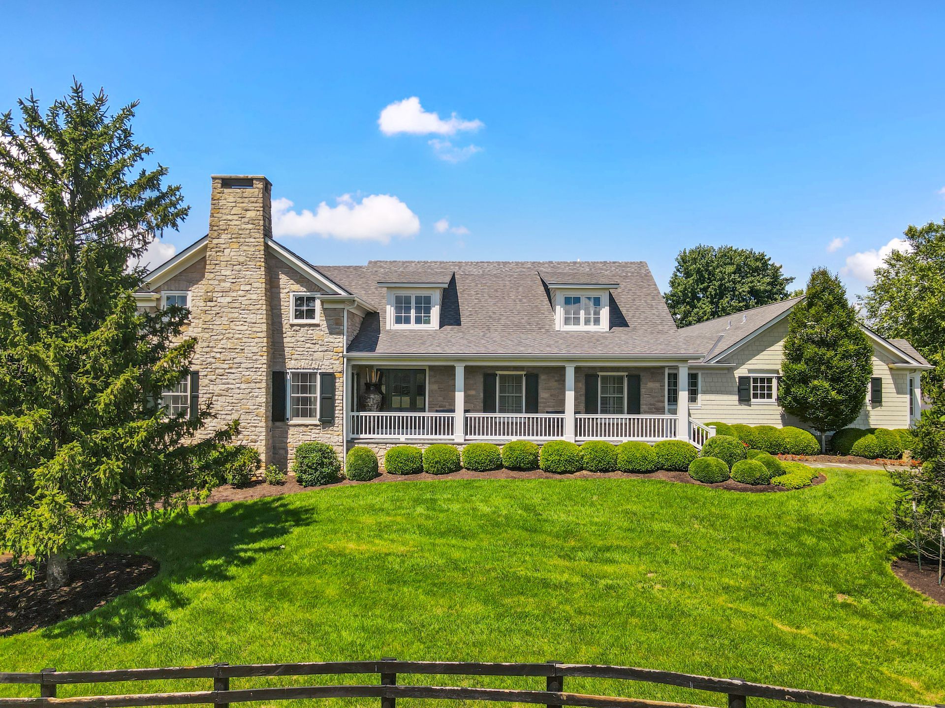 Stone and gray-sided house with a wraparound porch and chimney on a grassy hill under a blue sky.