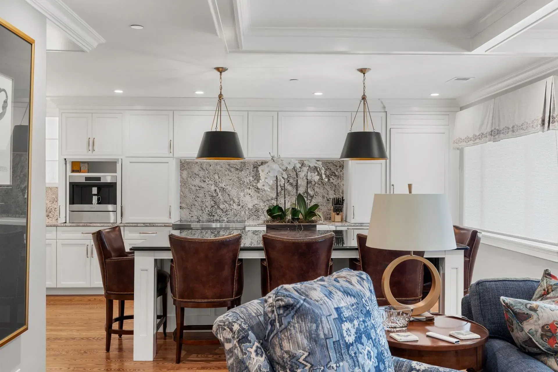 Kitchen with white cabinets, granite countertop, pendant lights, bar stools, and a floral armchair.