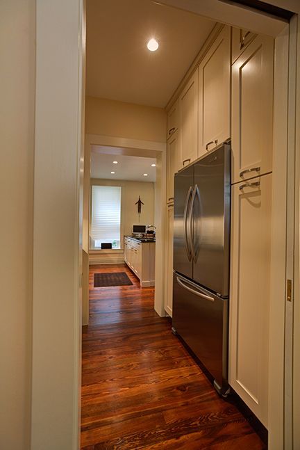 Narrow kitchen with refrigerator, wood floor, and cabinets; leads to another room.