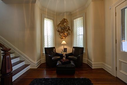 Entryway with bay windows, two chairs, a footstool, and stairs. Artwork hangs between windows.