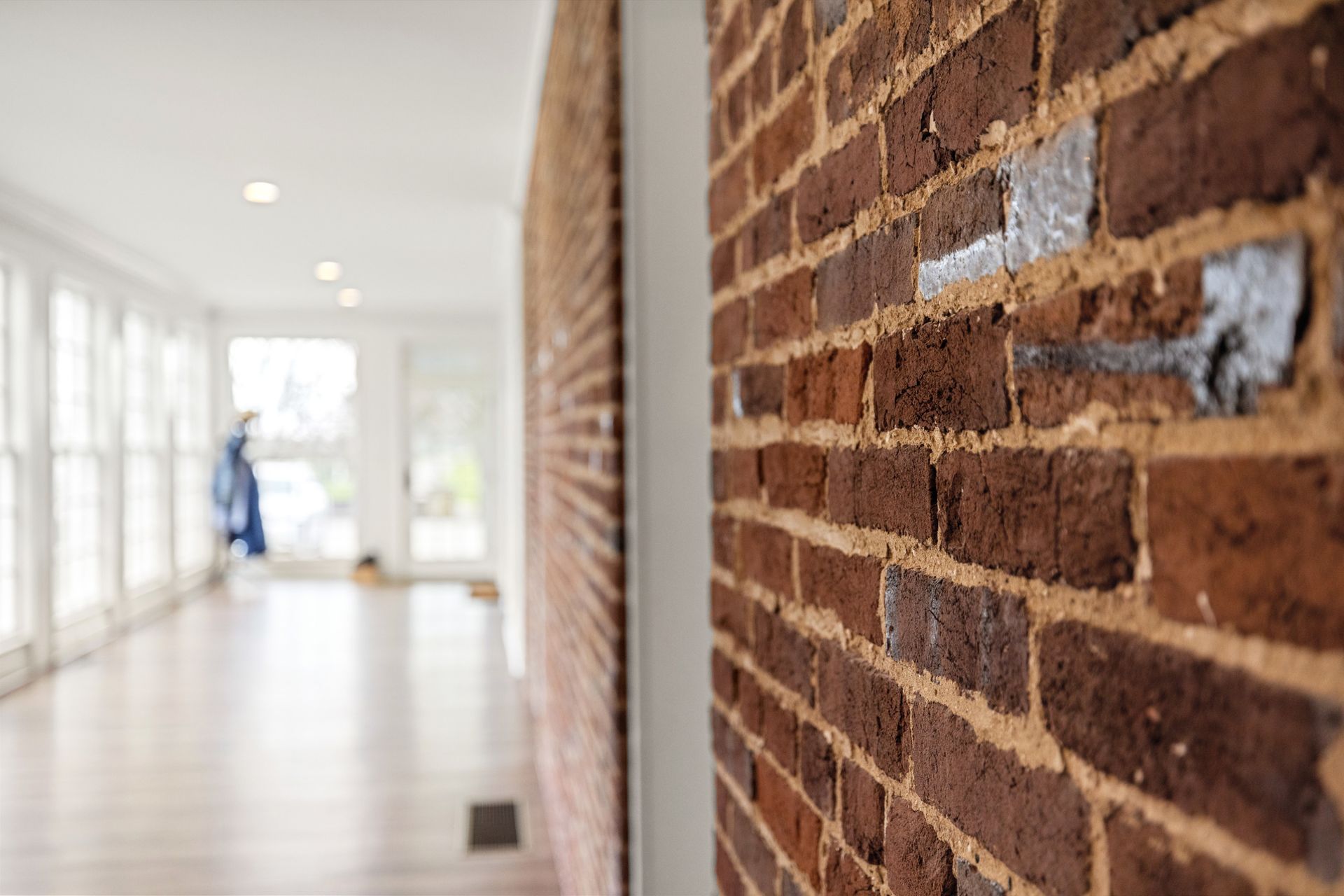 Brick wall in focus, leading to a blurred hallway with windows and a figure.