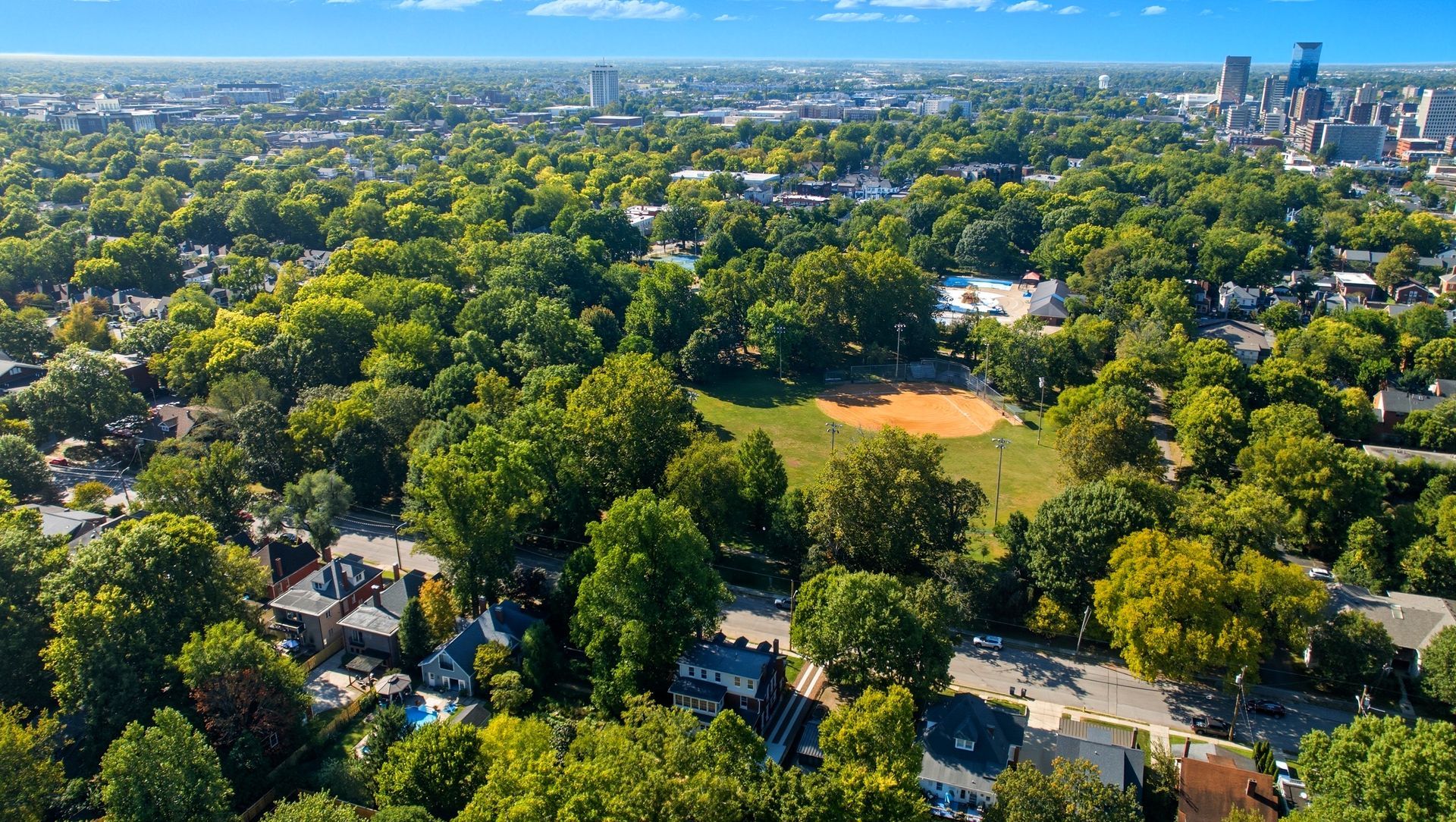 Aerial view of a lush green city park with a baseball field, surrounded by trees and residential buildings.