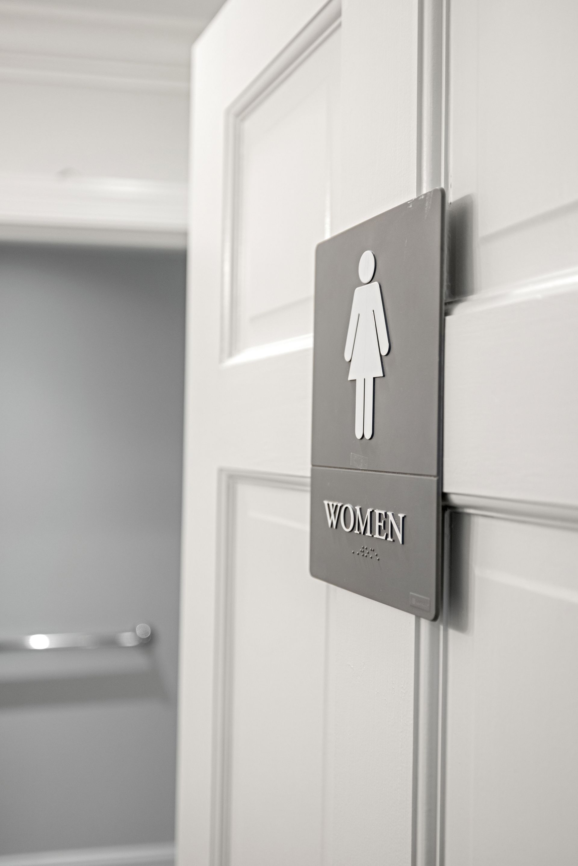 Gray and white women's restroom sign on a white door, with the open door revealing a gray interior and towel rack.
