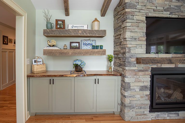Built-in shelving with wooden beams, gray cabinets, and a stone fireplace in a room with wood flooring.