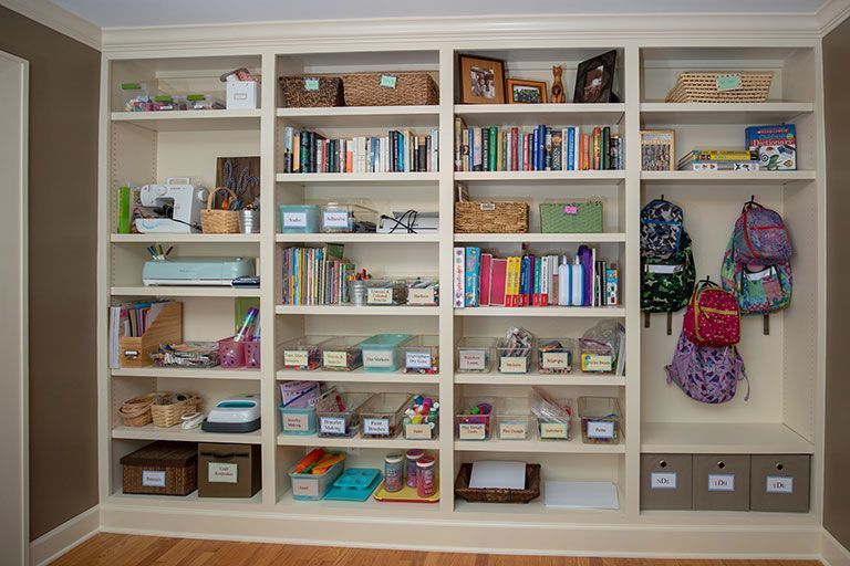 Built-in white bookshelf with books, craft supplies, and backpacks in a light brown room.
