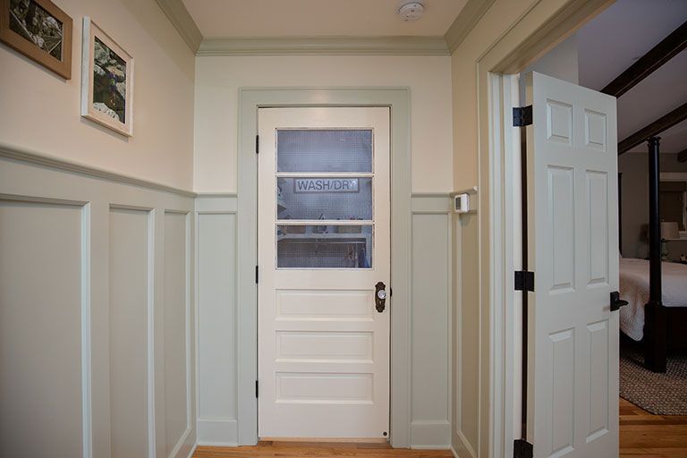 A hallway with light green wainscoting, a white door with a frosted glass panel, and an open door leading to a bedroom.