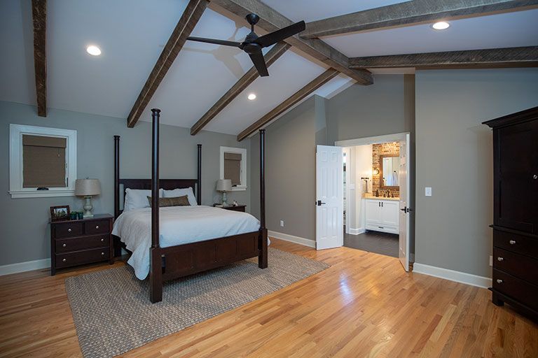 Bedroom with vaulted ceiling, wooden beams, four-poster bed, wood floors, and an open door to a bathroom.