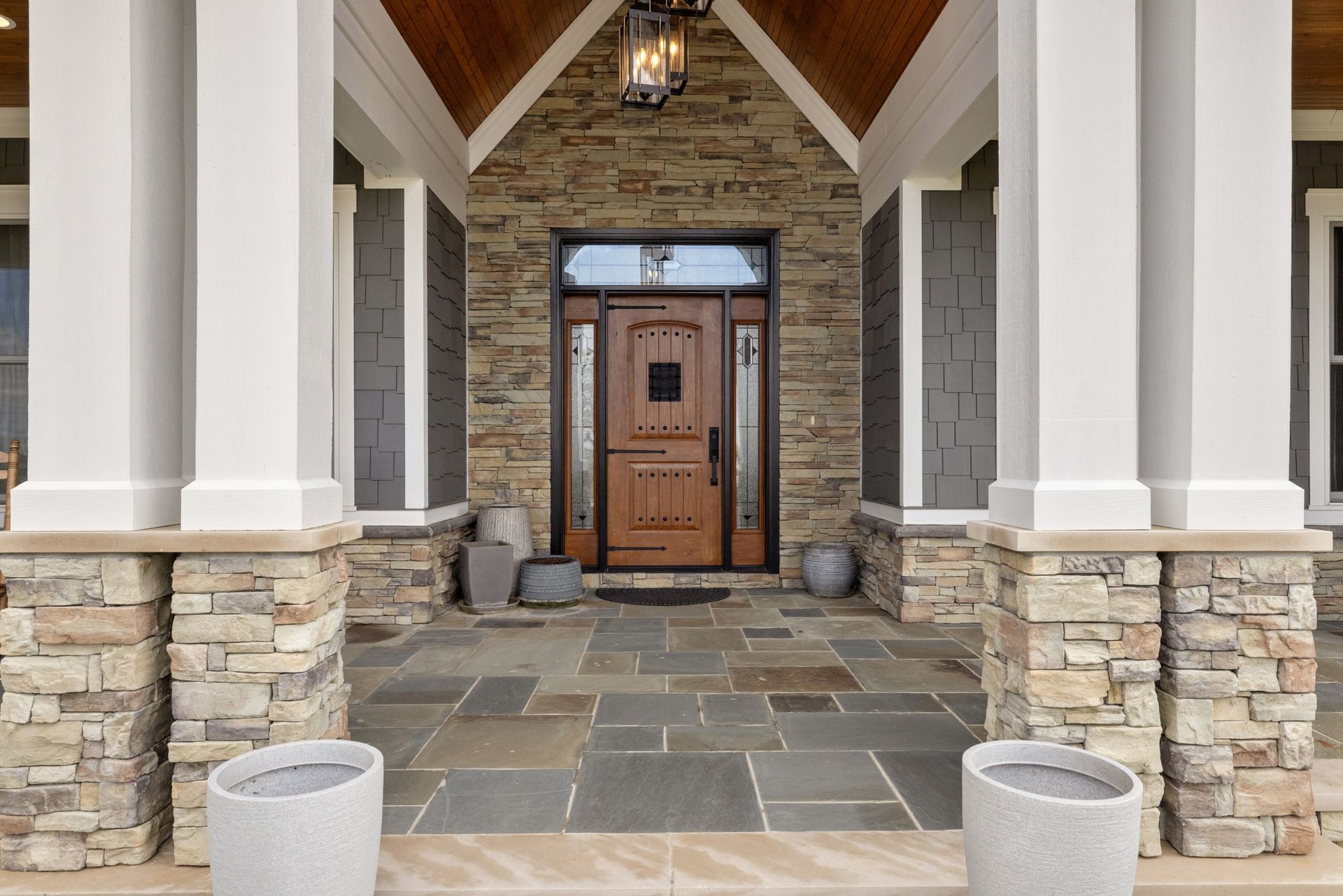 Front entrance with stone columns, brown door, gray stone and slate flooring, and gray planters.