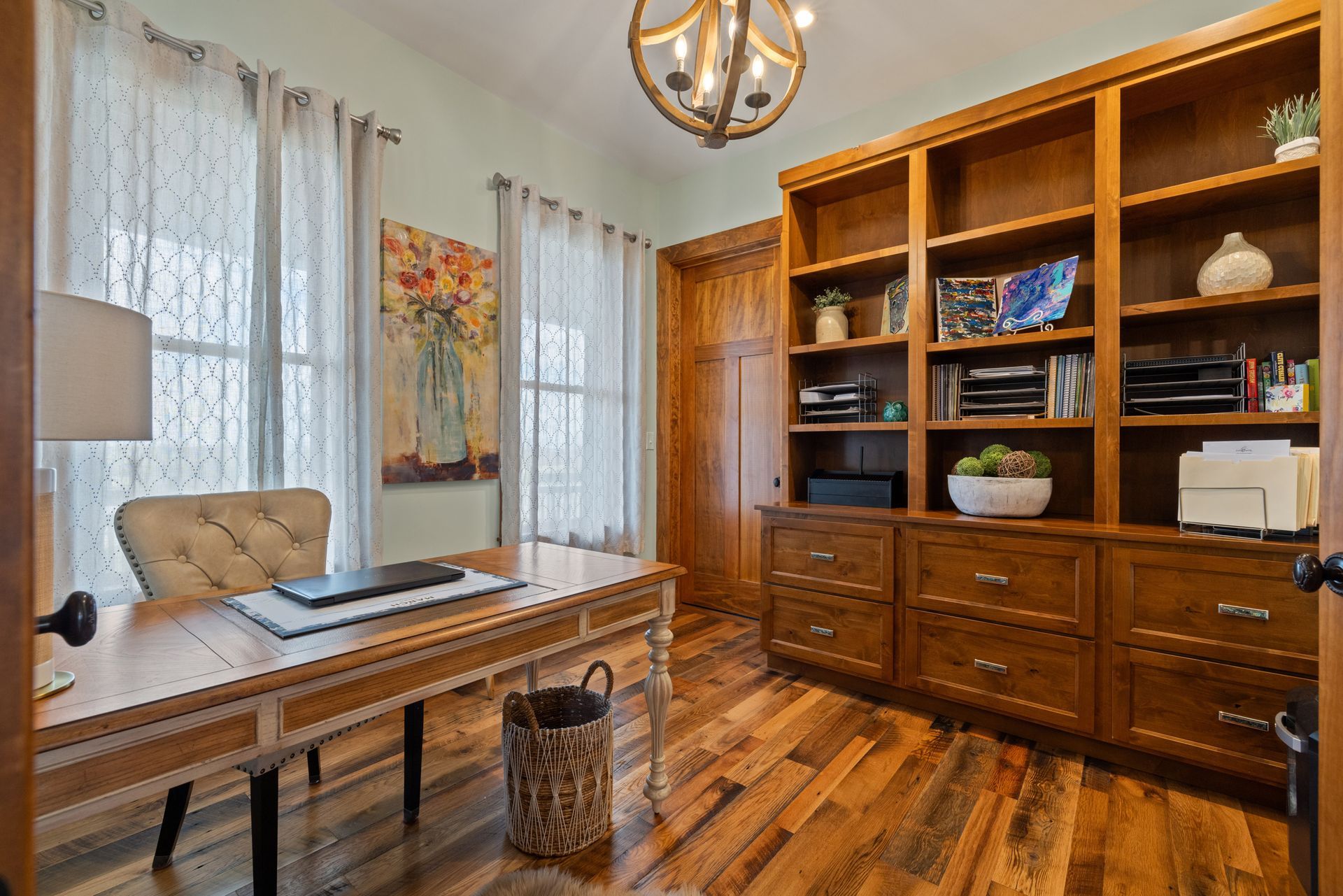 Home office with wooden desk, built-in bookcase, art, and arched chandelier.