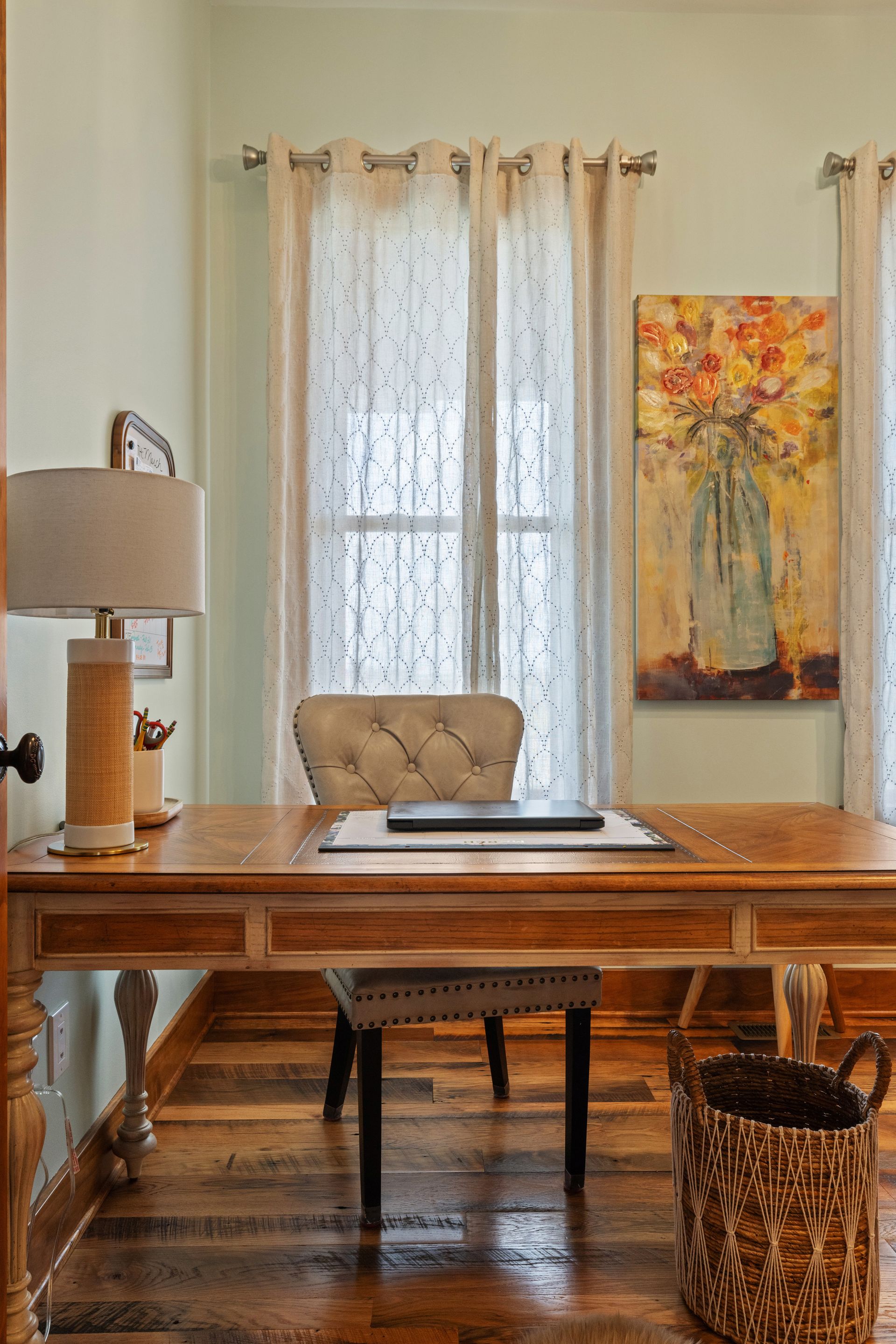 Desk in light green room with window, sheer curtains, and colorful floral art.