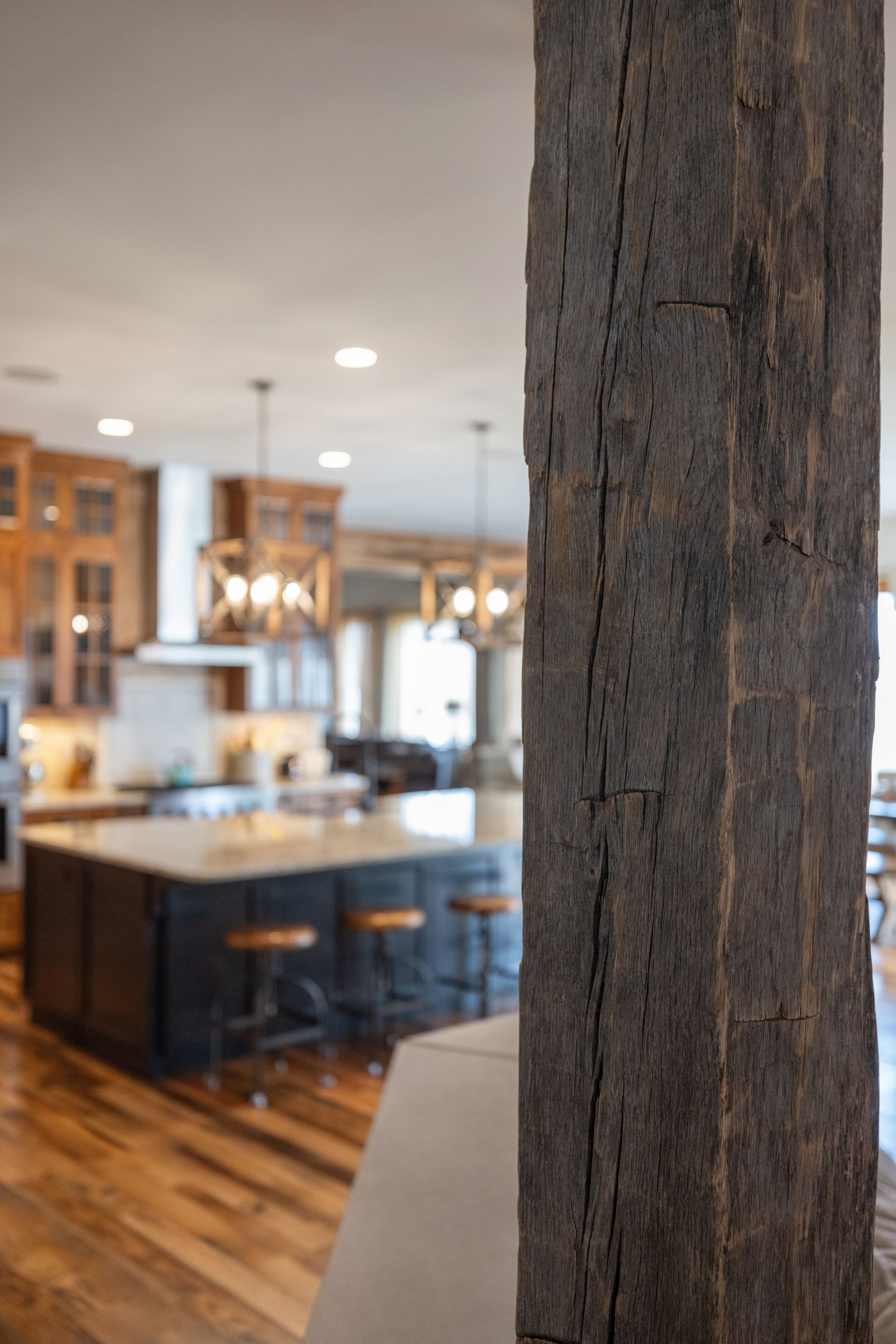 Close-up of a weathered, dark wooden beam in a kitchen. In the background is a kitchen island and cabinetry.