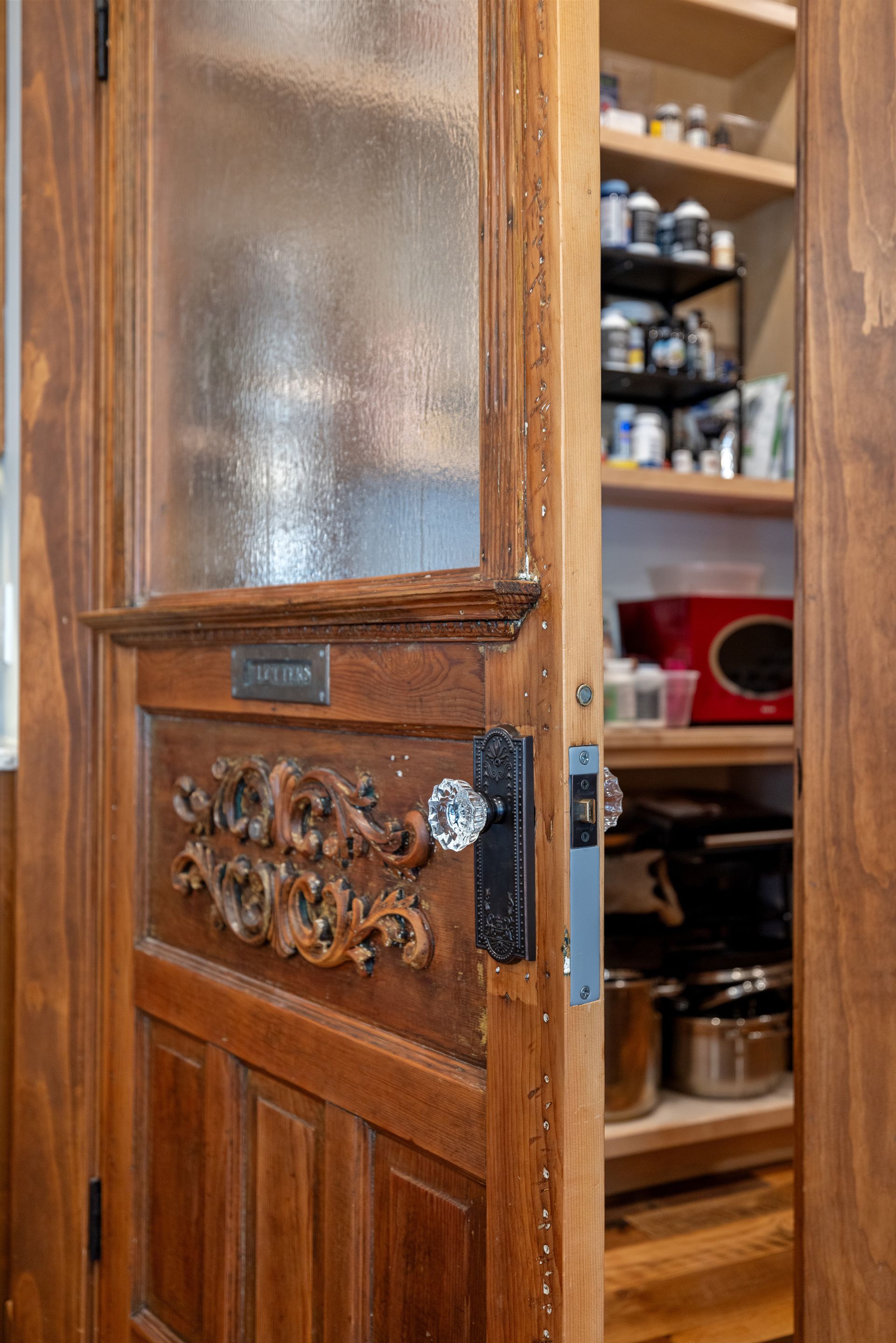 Open pantry door, wood with ornate carvings and frosted glass, reveals shelves with food items.