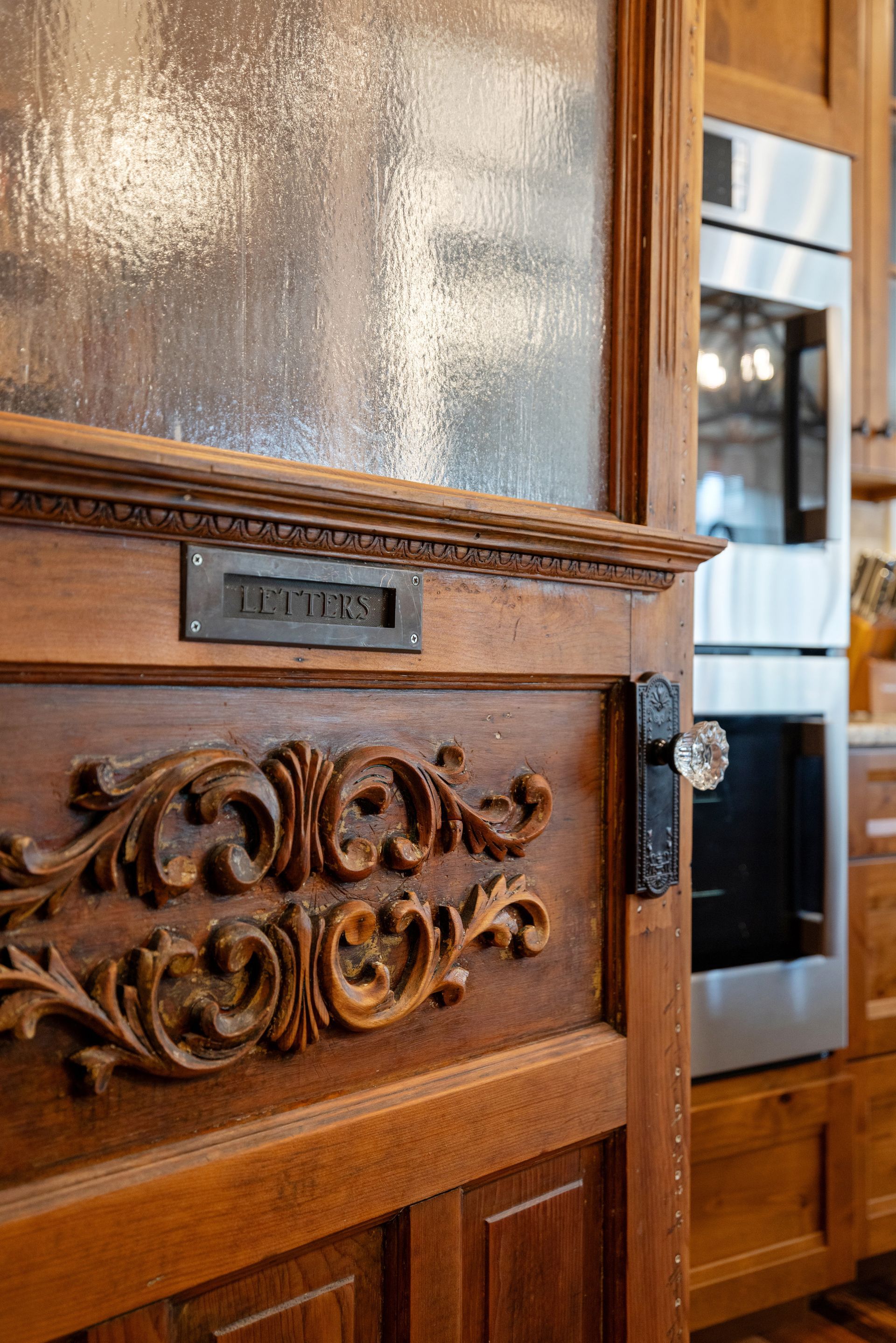 Ornate wooden door with glass panel and carved detailing, revealing kitchen with oven.