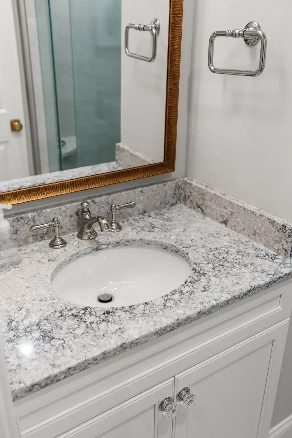White bathroom vanity with oval sink, granite countertop, gold-framed mirror, chrome fixtures, and crystal knobs.