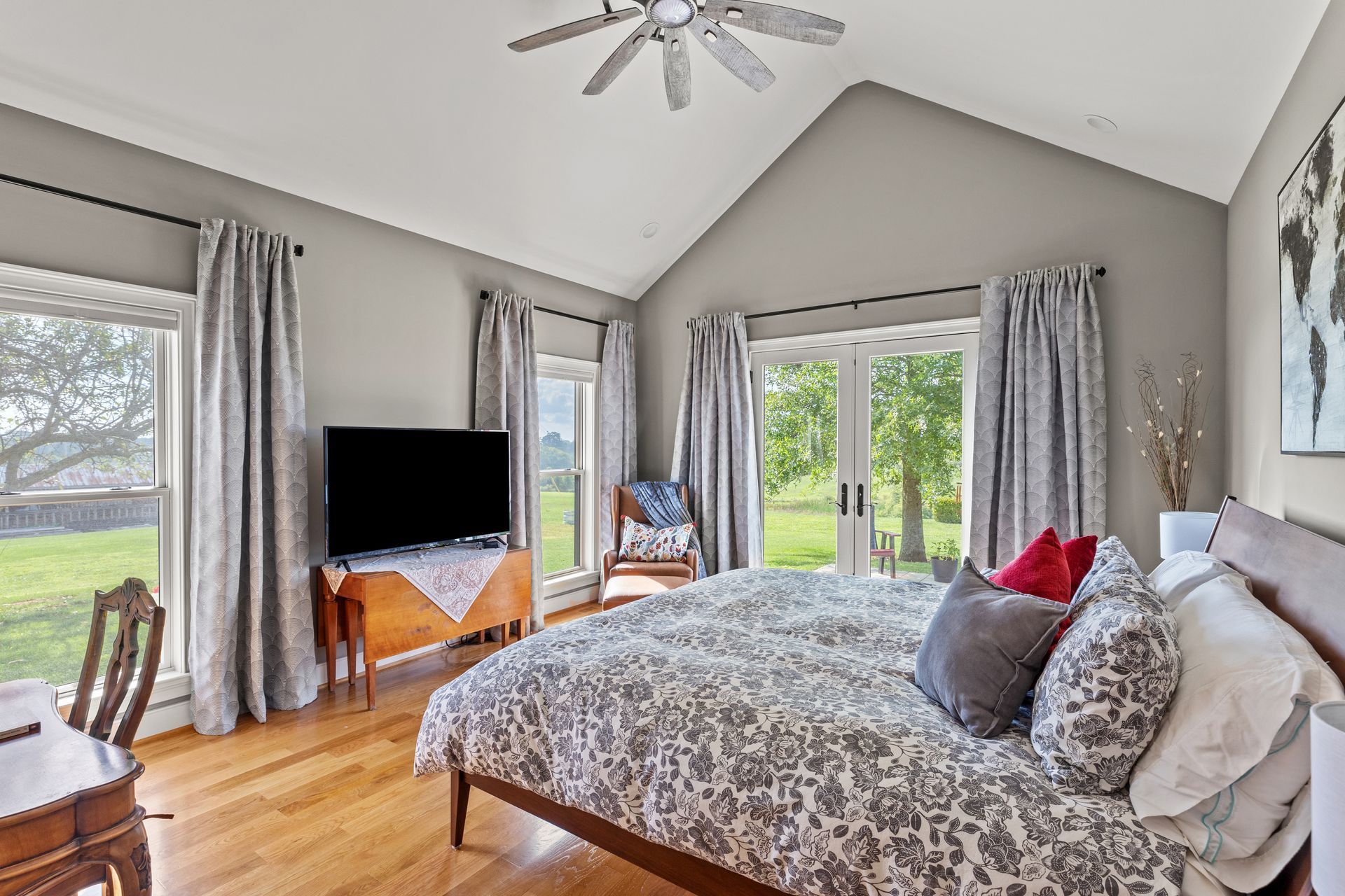 Bedroom with a bed, windows, TV, and curtains; light gray walls, and hardwood floors.
