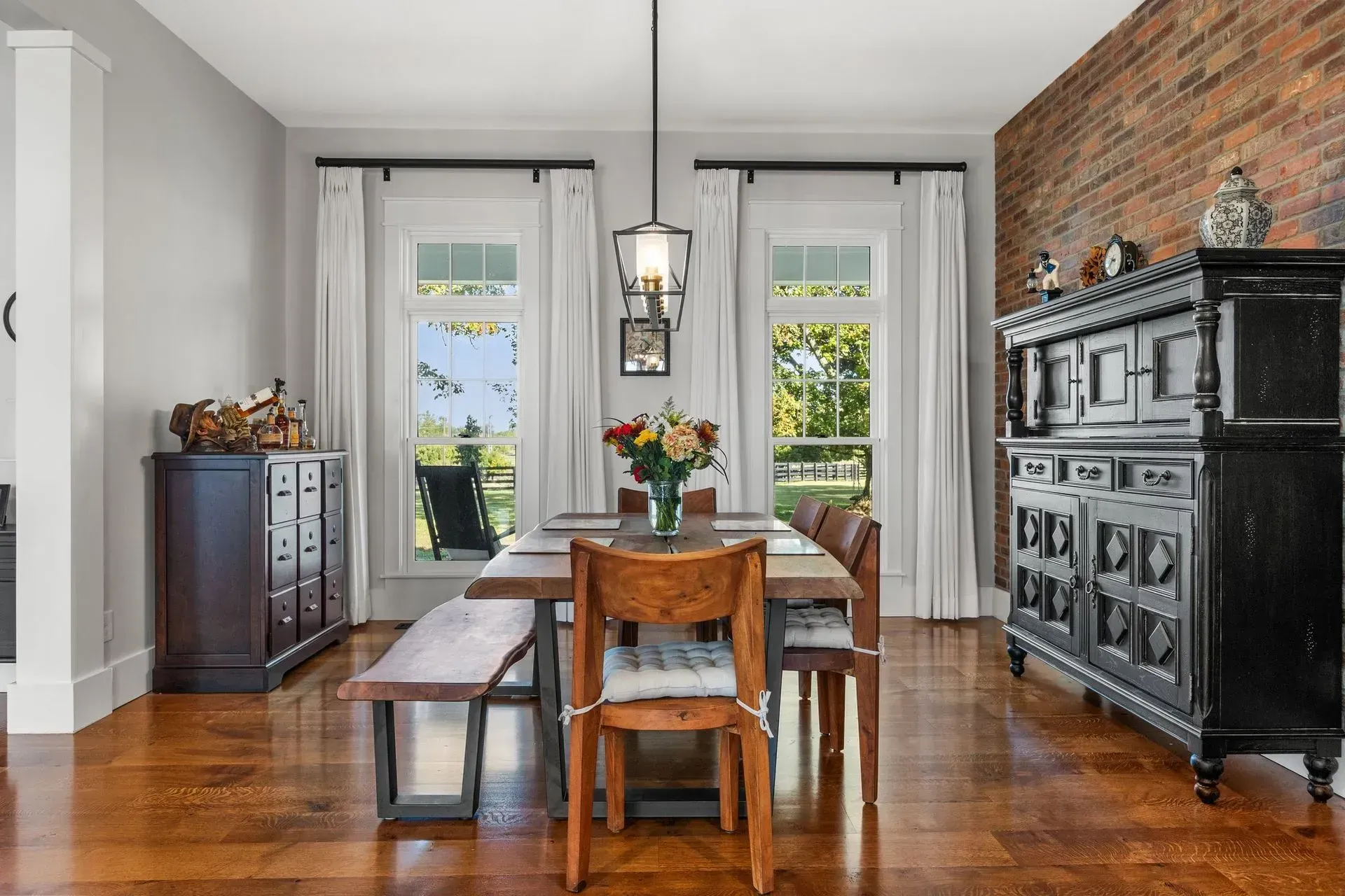 Dining room with a wooden table, chairs, and a bench, featuring windows with white curtains, brick wall, and hardwood floor.