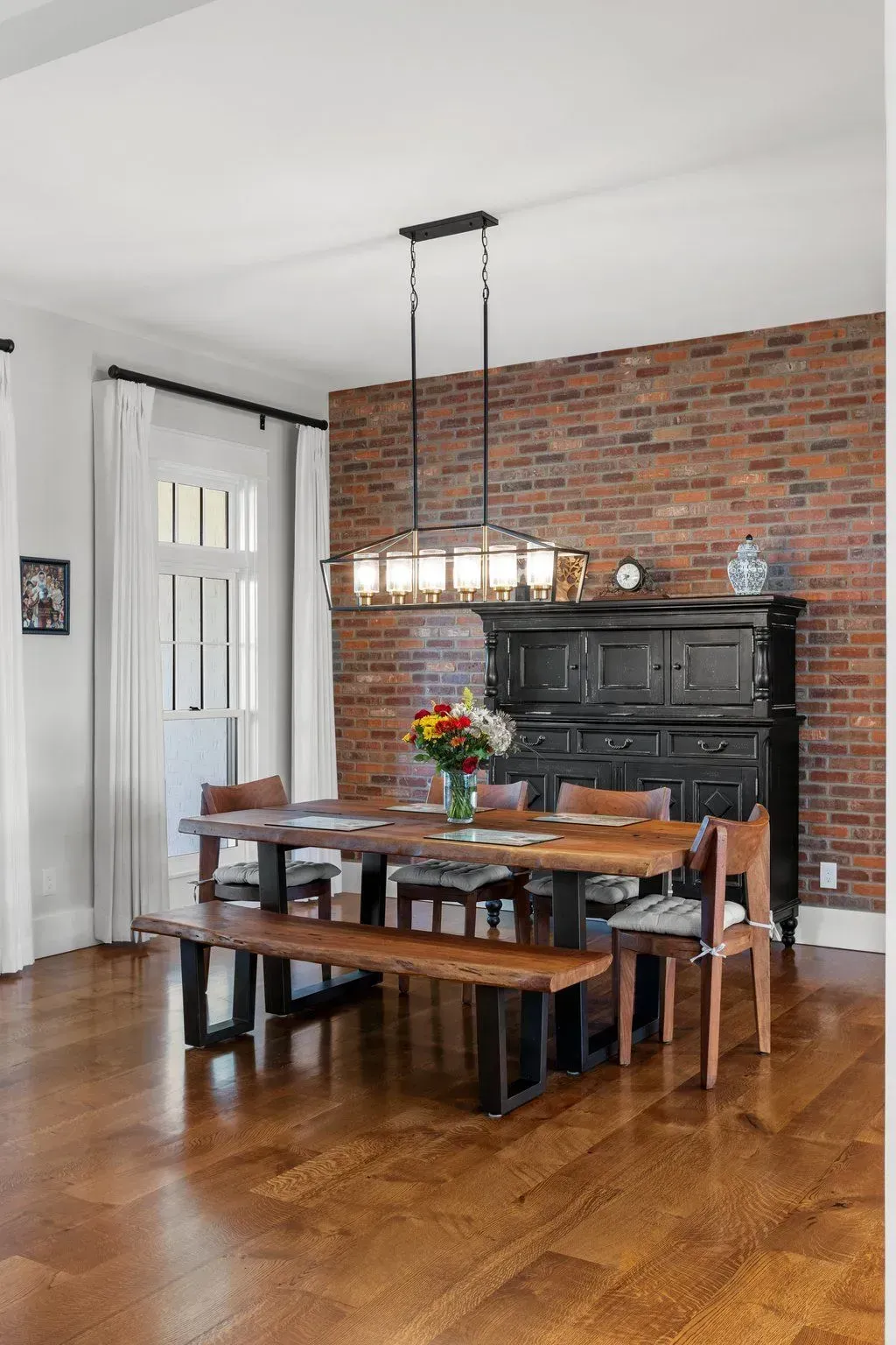 Dining room: Wooden table with benches and chairs, brick wall, black cabinet, chandelier.