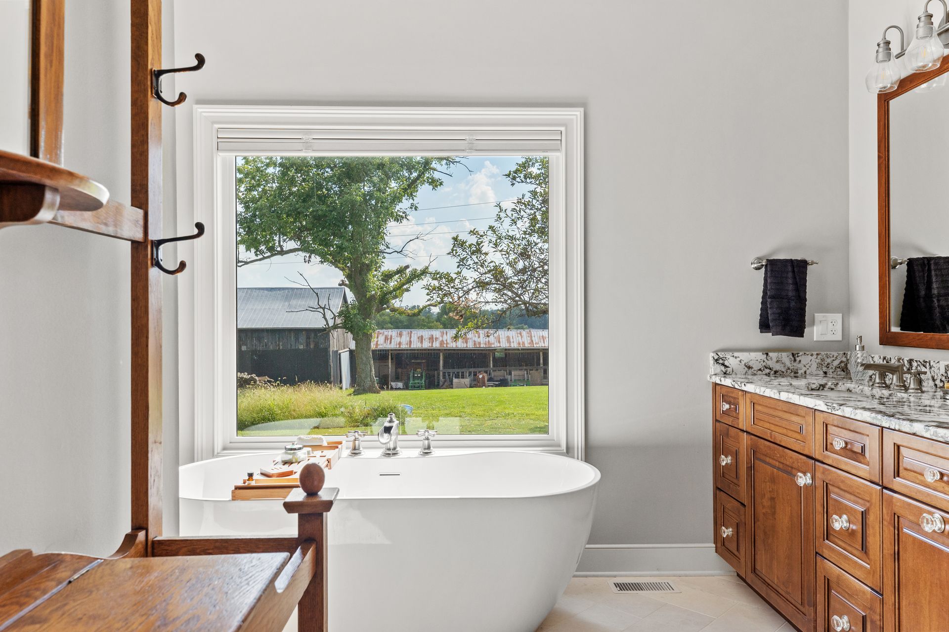 Bathroom with a white tub in front of a window overlooking a yard. Wooden cabinets and a vanity mirror.