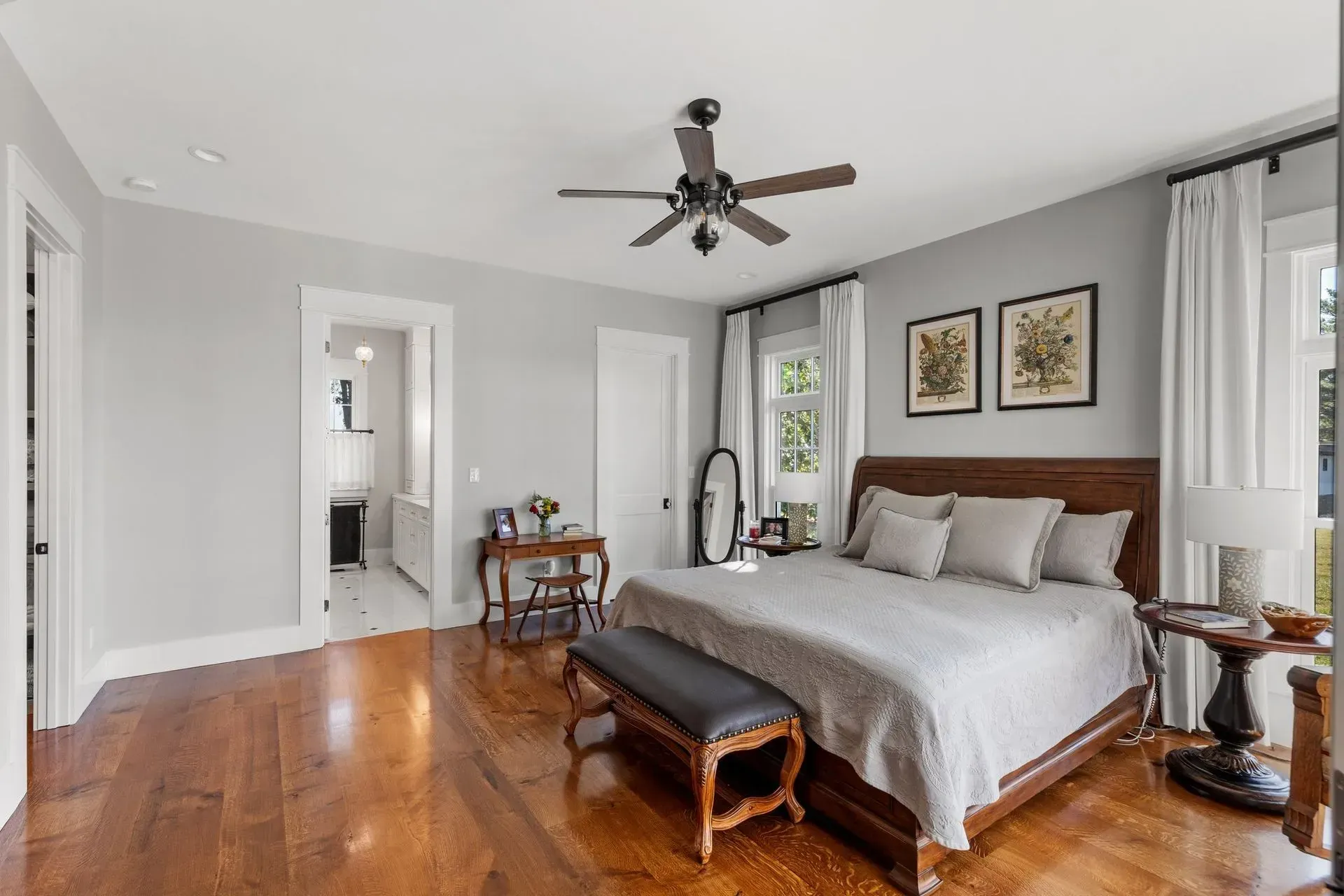 Bedroom with hardwood floor, bed, and access to a bathroom. Light gray walls, white trim, and a ceiling fan.