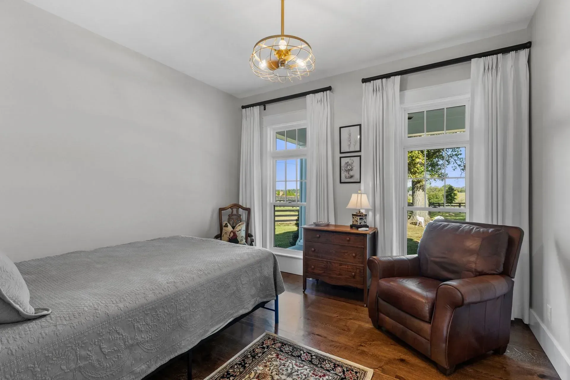 Bedroom with a bed, leather armchair, and chest of drawers. Two windows with white curtains.