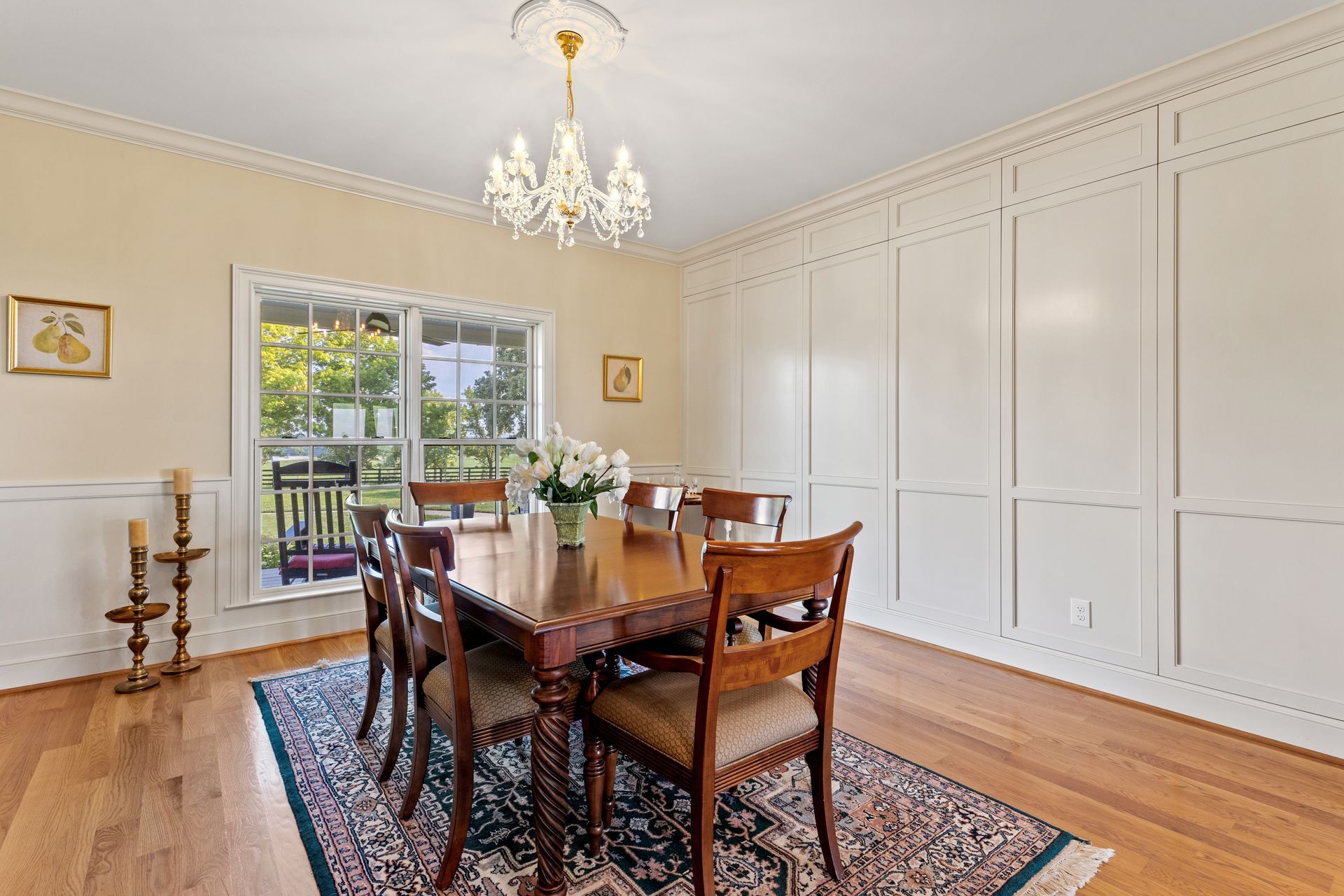 Dining room with wooden table, chairs, chandelier, and rug, set against a white paneled wall and window.