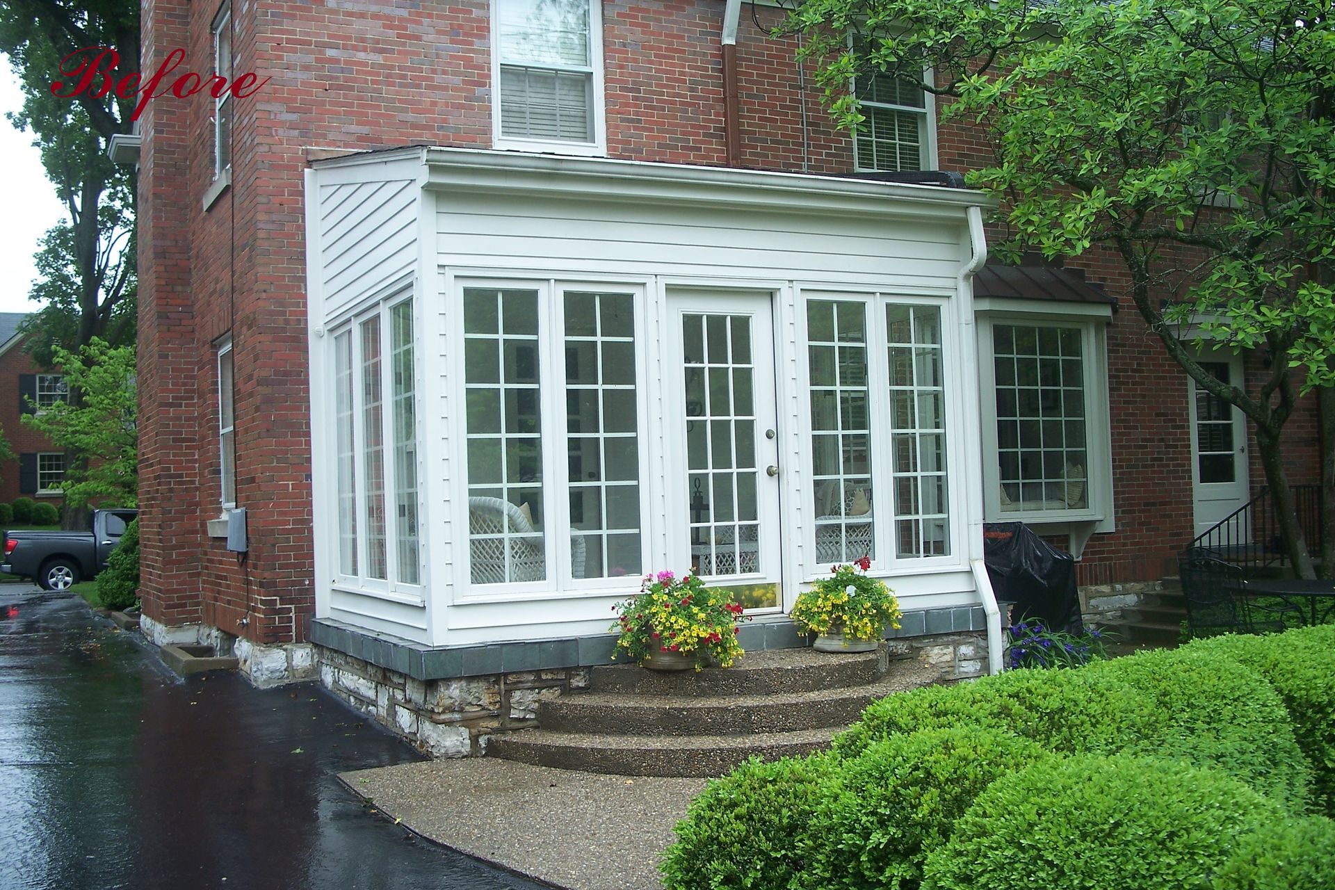 White sunroom attached to a brick house, with a stone stair entrance and green bushes.