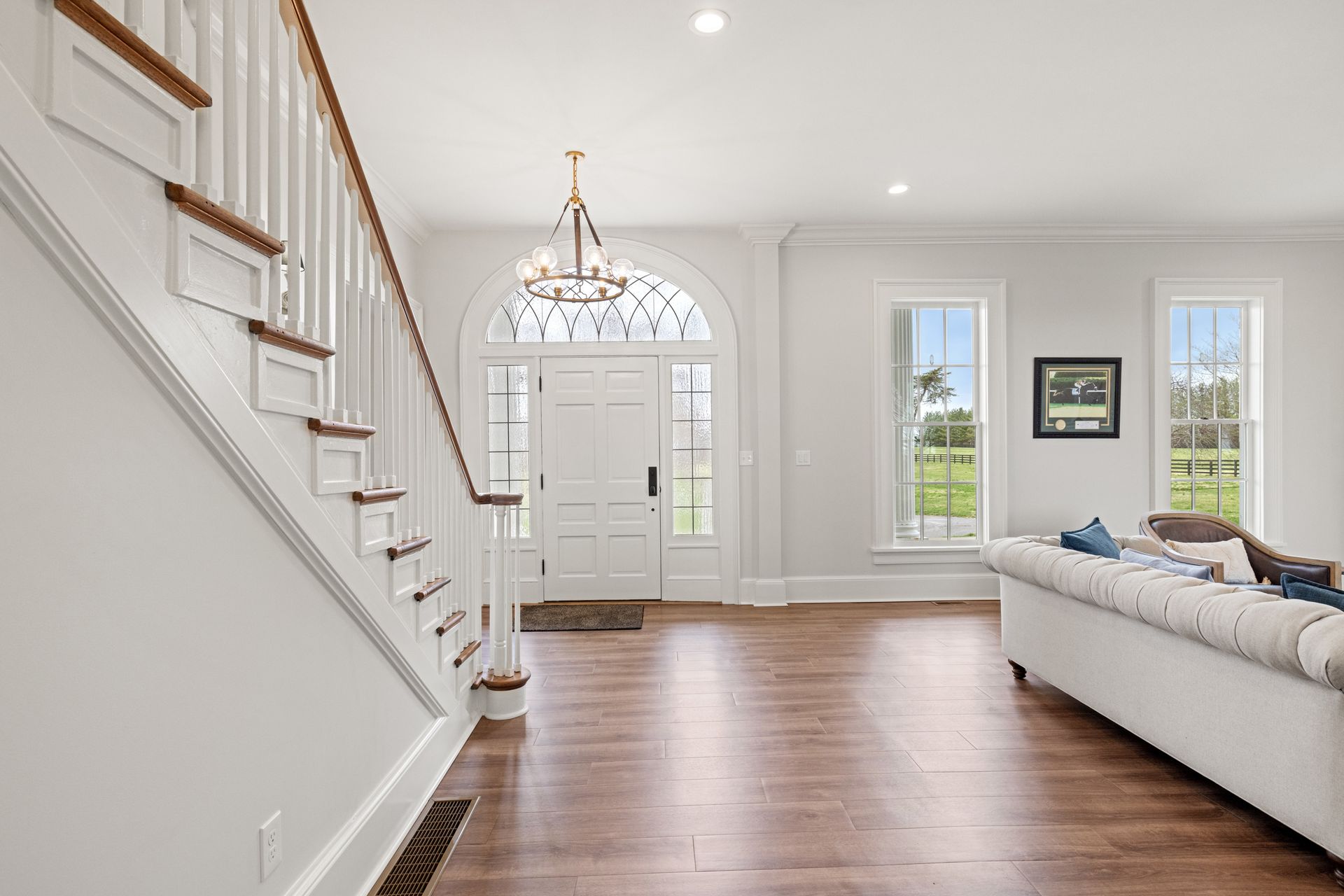 Bright interior: entry hall with stairs, front door, hardwood floors, chandelier, and a white couch.