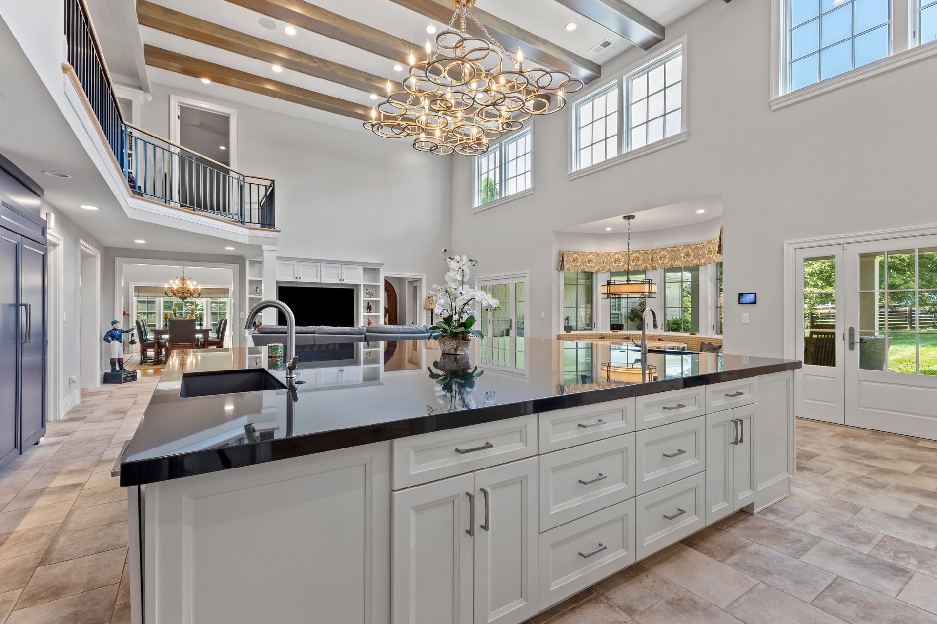 Spacious white kitchen with large island, black countertop, and chandelier; leads to dining and outdoor spaces.