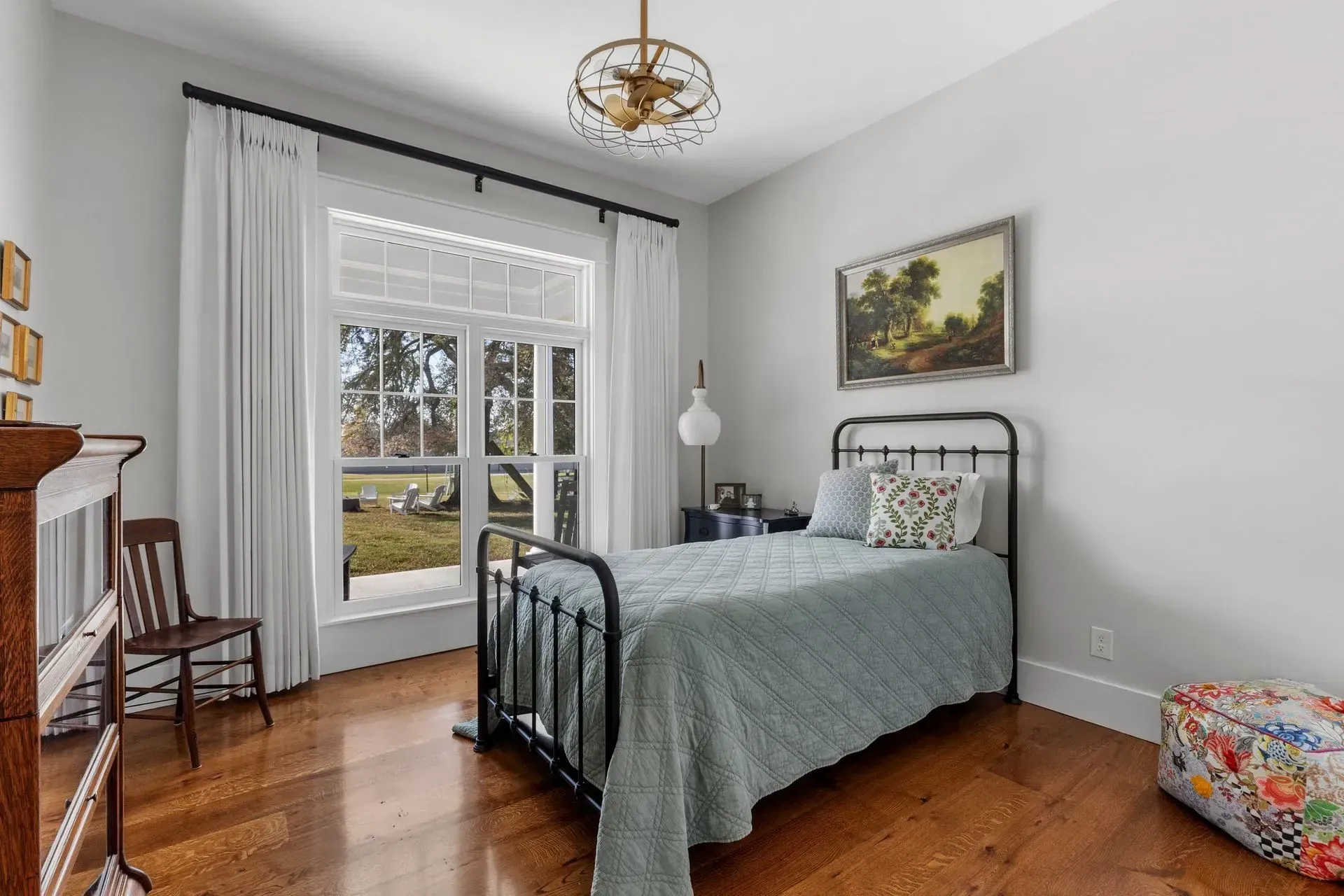 Bedroom with a metal bed, floral ottoman, large window with white curtains, and a landscape painting.