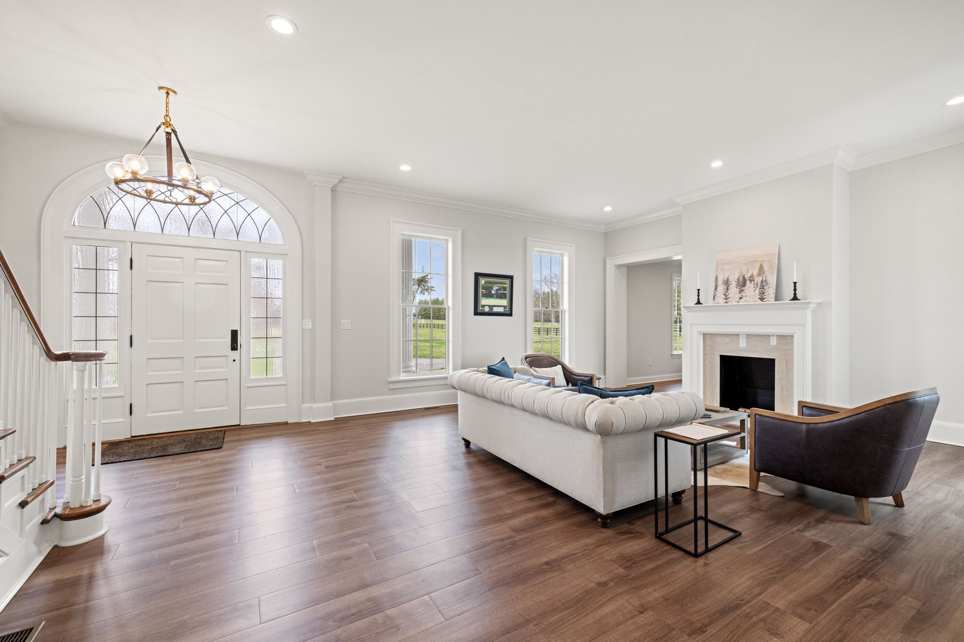 Spacious, bright living room with dark wood floor, fireplace, and large windows. White walls, couch, and chandelier.