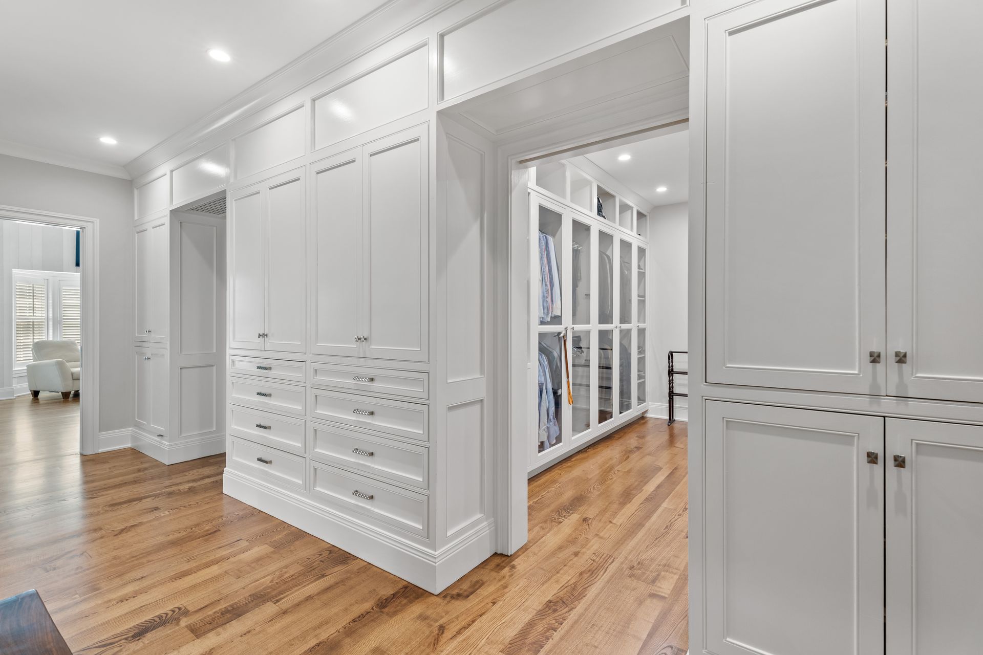 A spacious, white walk-in closet with drawers and cabinets, hardwood floors, and a view into another closet.