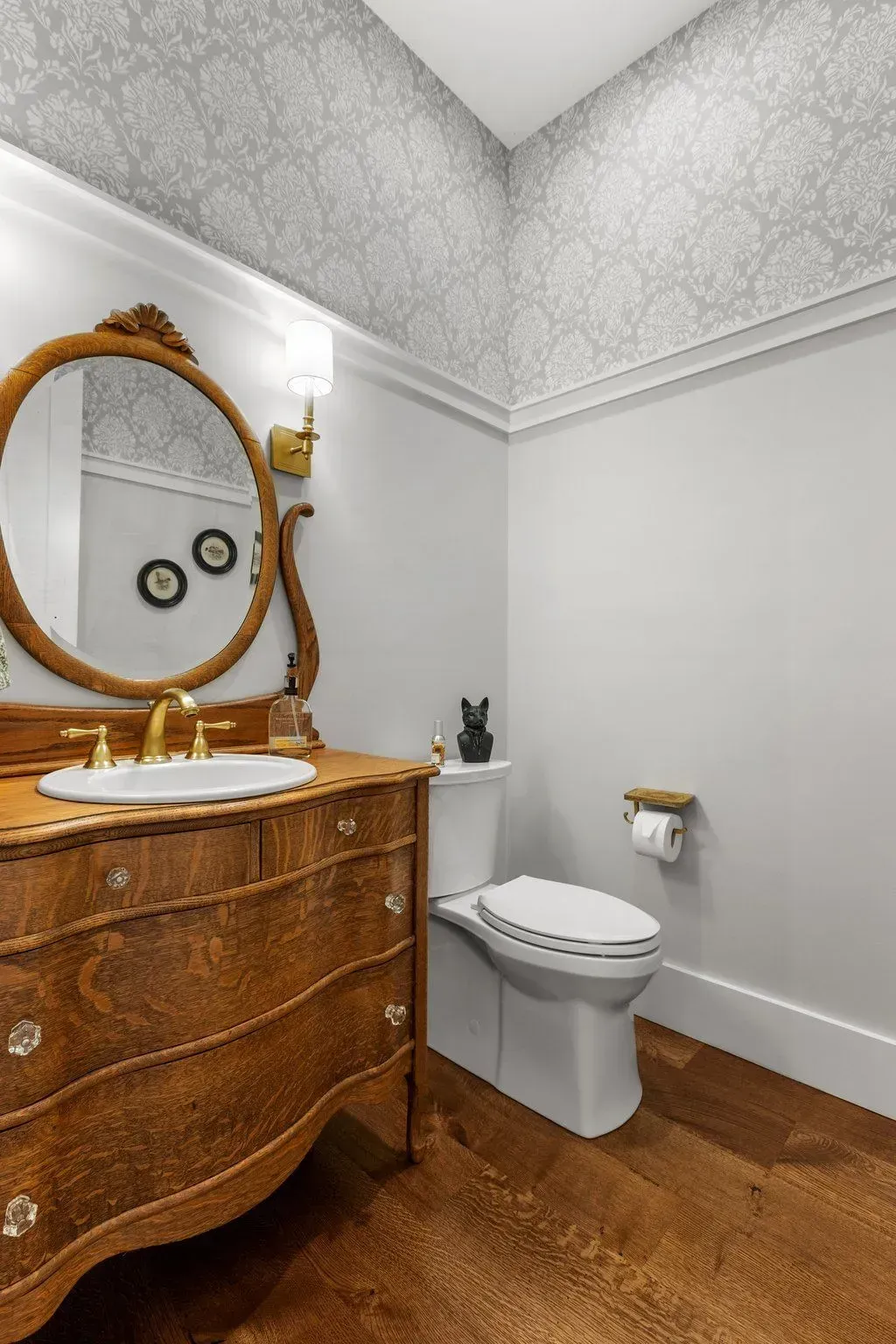 A small bathroom with a wood vanity, toilet, floral wallpaper, and hardwood floor.