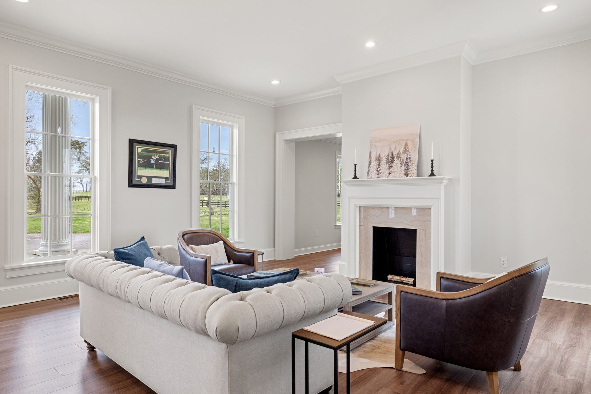 Spacious living room with white walls, fireplace, large windows, a cream tufted sofa, and two brown leather chairs.
