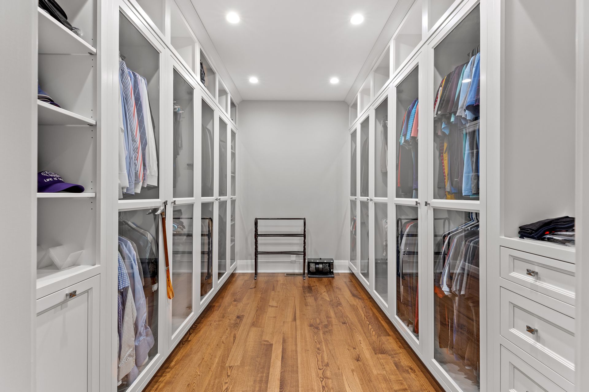 White walk-in closet with glass-door cabinets, hardwood floor, clothes hanging, and shoe rack.