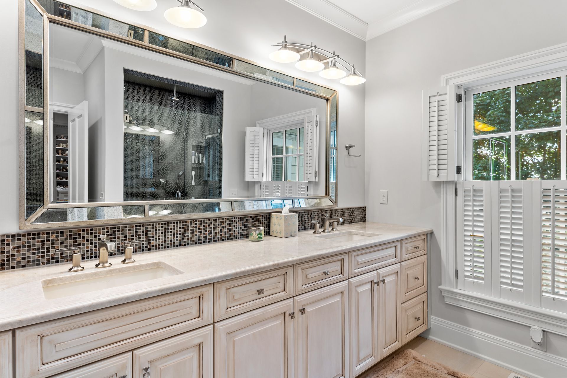 Bathroom with white cabinets, marble countertop, large mirror, and a window with shutters.