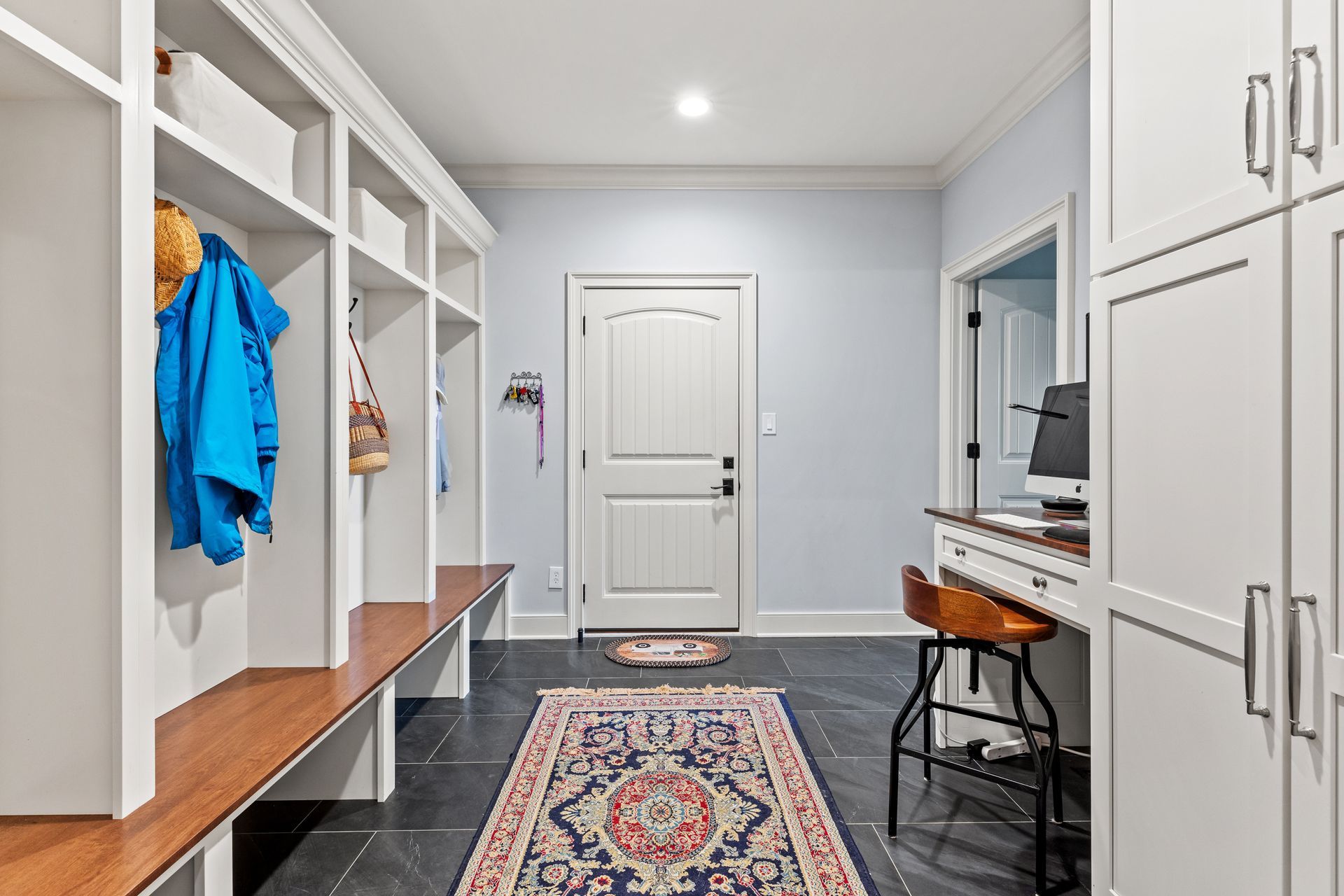 Mudroom with lockers, bench, rug, desk, and door; light blue walls, white cabinets, and slate floor.