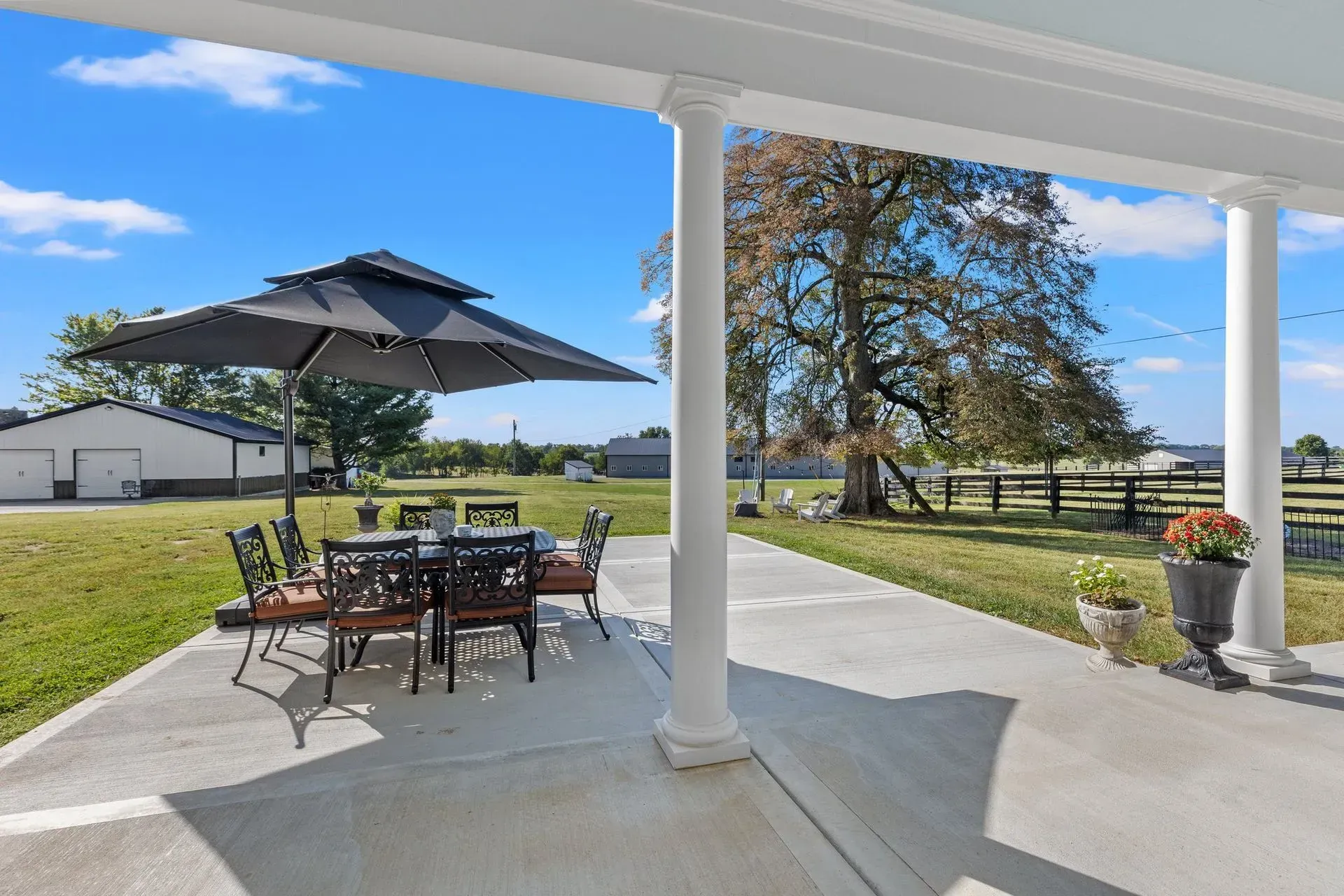 Patio with outdoor furniture under a black umbrella, overlooking a grassy field and barns.