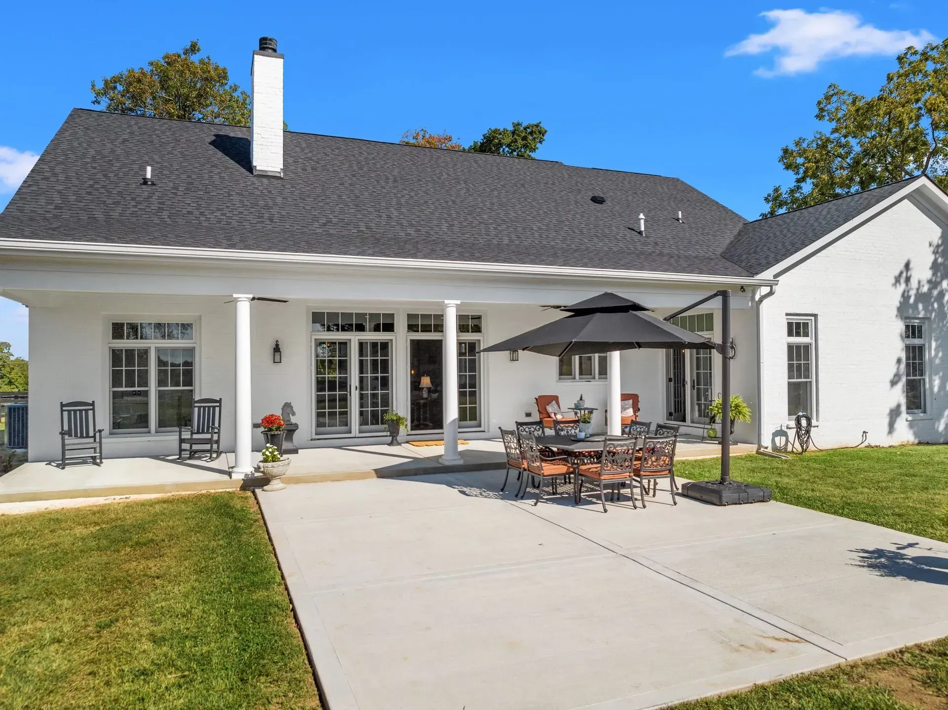 Back patio of white brick house with outdoor dining set and umbrella on a sunny day.