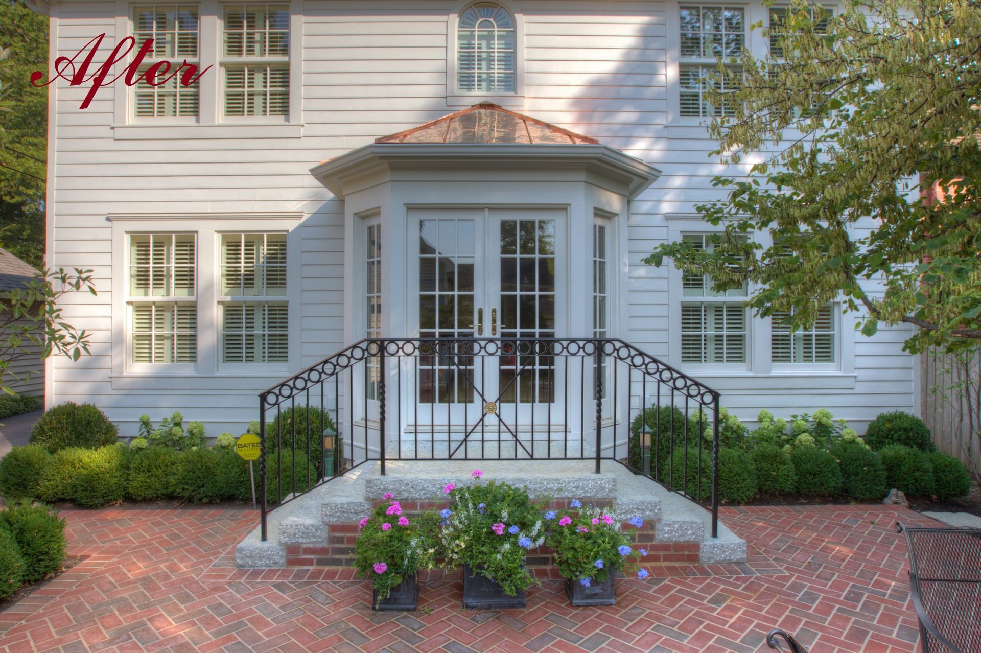White house with a brick patio, black railing, and flower planters.