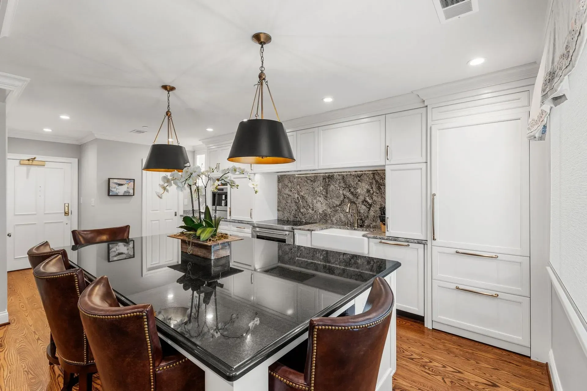 Luxury kitchen with black granite island, pendant lights, white cabinets, brown leather chairs, and stainless steel appliances.