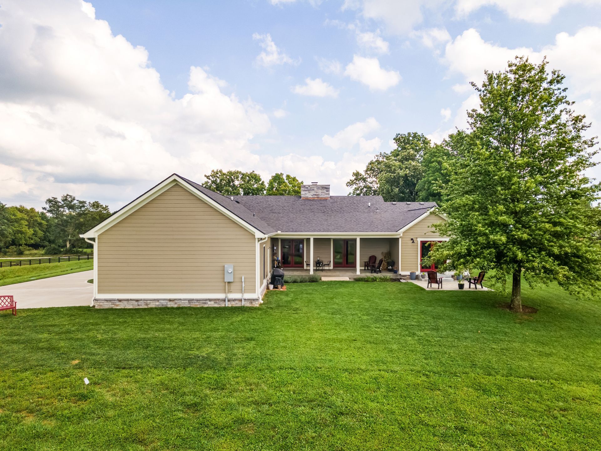 Tan ranch-style house with dark roof and porch, green lawn and trees under a partly cloudy sky.