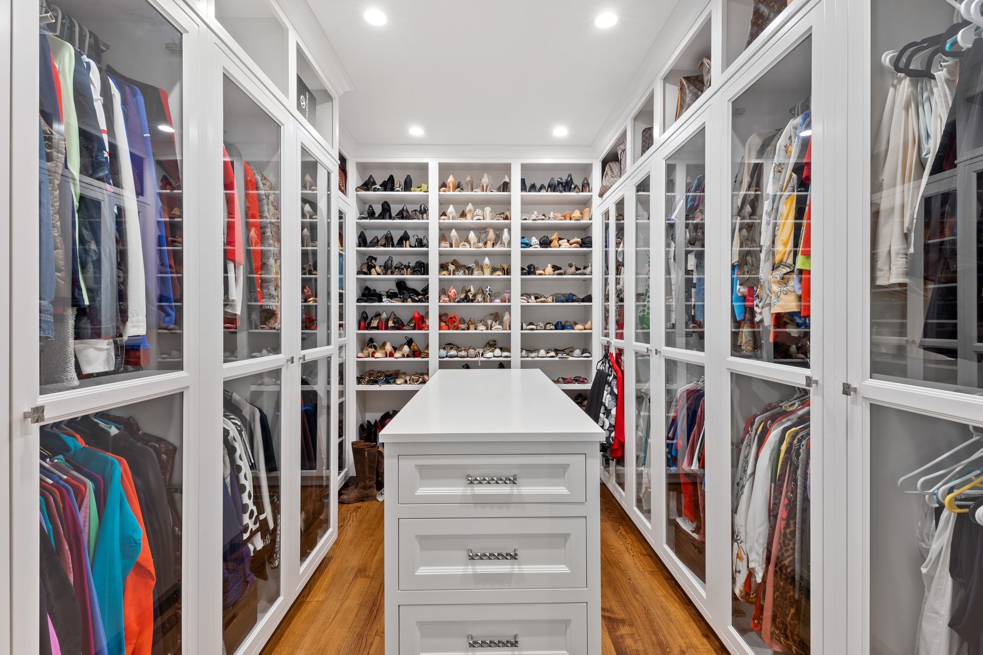 Walk-in closet with white cabinets, glass doors, and a central island; shoes and clothing are displayed.