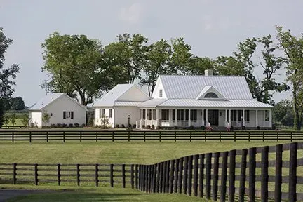 White farmhouse with metal roof, porch, and outbuilding behind a wooden fence on a grassy field.