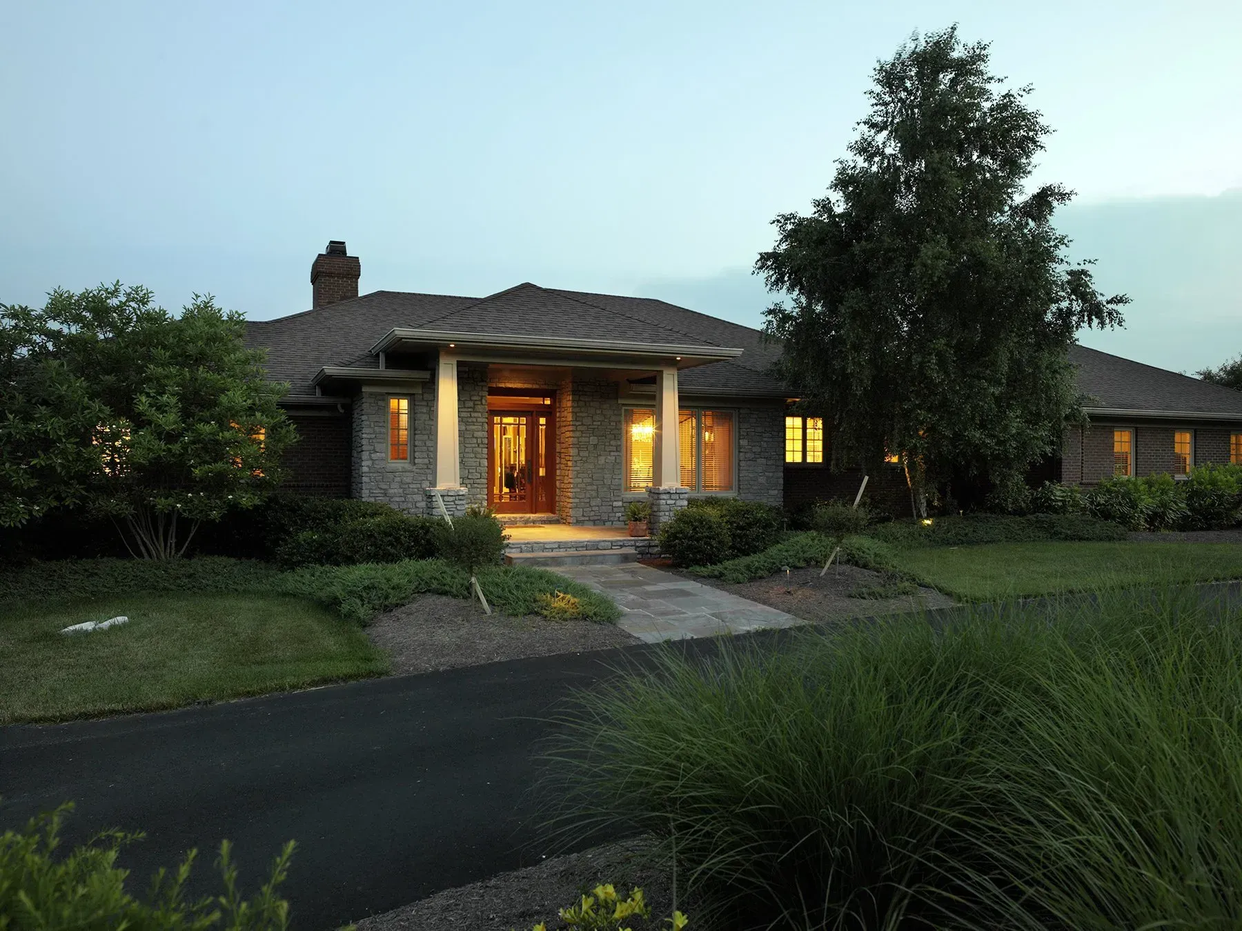 Stone home at dusk with illuminated windows, a walkway, and lush landscaping.