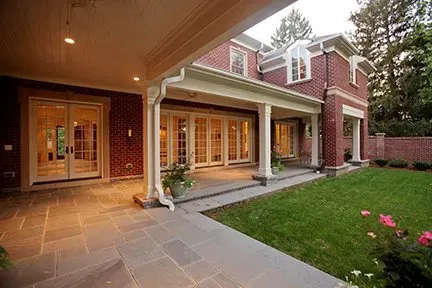 Covered patio of a red brick house with French doors, a green lawn, and pink flowers.