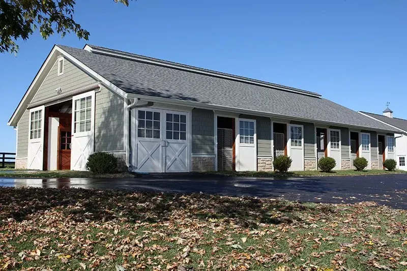 A long, gray horse stable with open white doors under a clear blue sky; autumn leaves on the ground.