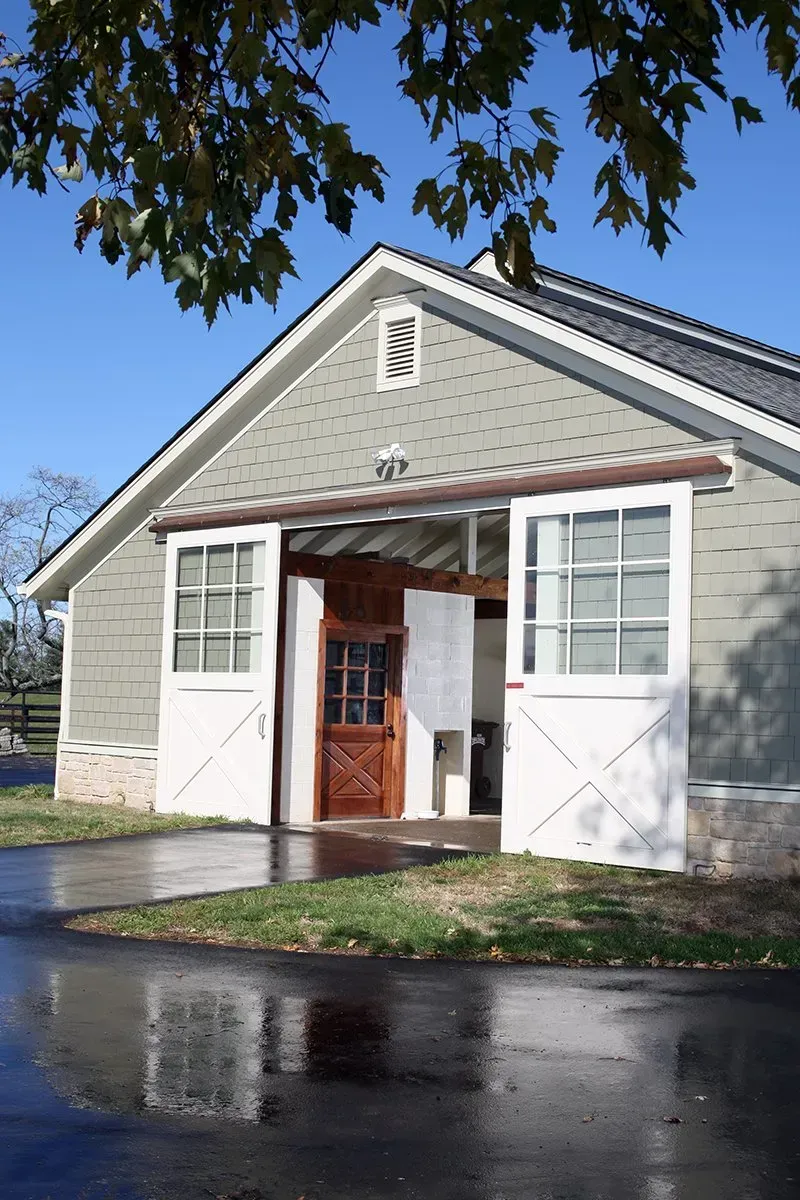 Green barn with white sliding doors and a wooden door, set on a paved area with reflection.
