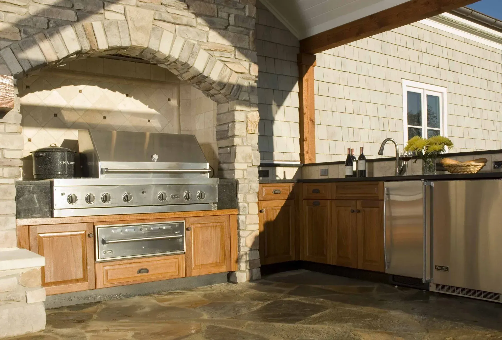 Outdoor kitchen with stone arch, stainless steel grill and appliances, wooden cabinets.