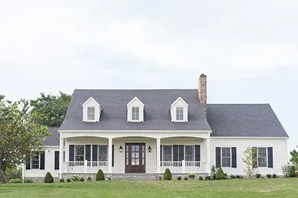 White farmhouse with dark roof, porch, black shutters, and dormers on a grassy lawn.