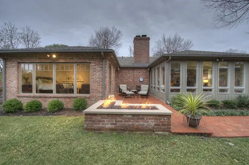 Backyard with brick patio, fire pit, seating, and house with large windows; green lawn, overcast sky.